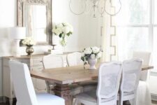 a chamring French country dining room with a neutral sideboard, a stained vintage table, white chairs, a lovely chandelier and a mirror in a wooden frame