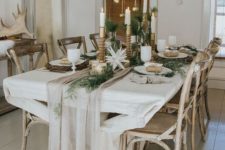 a chic and neutral winter tablescape with a neutral runner, greenery, paper stars candles in various candleholders and ornaments hanging over the table