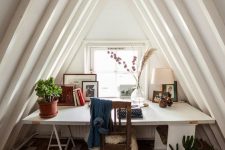 a cool attic home office with a desk by the window, a vintage chair and a reclaimed wood floor, potted greenery and artworks