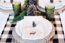 a cozy winter table with a plaid runner, wood slice placemats, large pinecones, green glasses and lots of candles