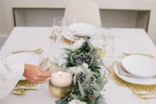 a glam winter tablescape with gold placemats, an evergreen, white bloom and snowy pinecone runner, candles and gold cutlery