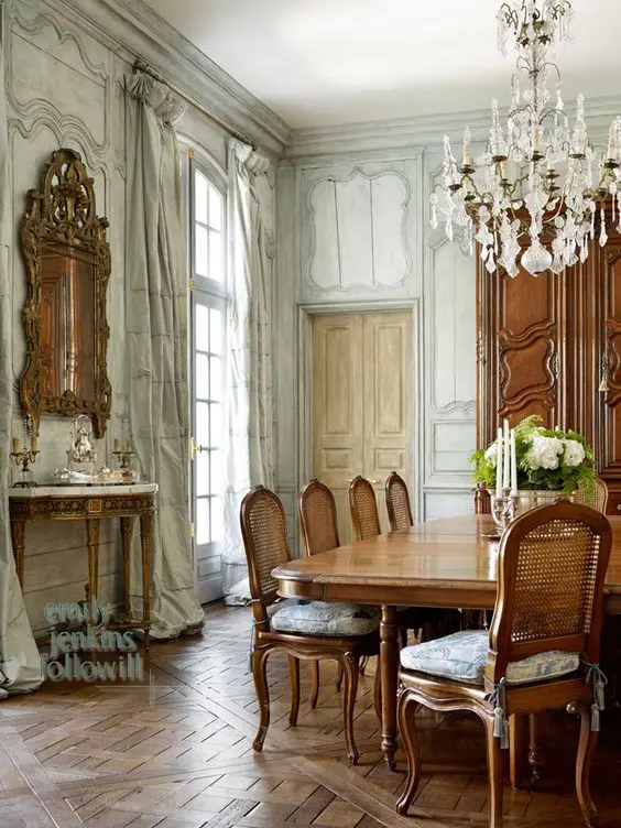 A jaw dropping French chic dining room with paneling, a chic console table, a large stained table and chairs, a crystal chandelier