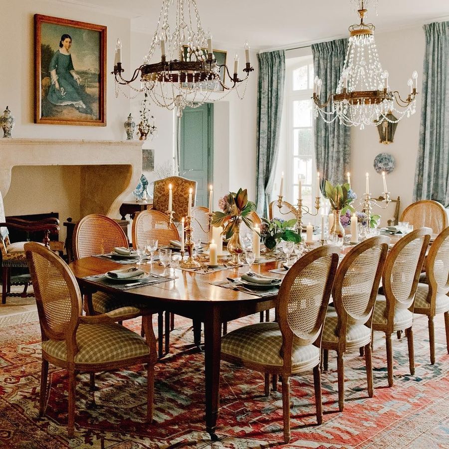 A jaw dropping vintage French dining room with a non working fireplace, a large stained table and vintage chairs, crystal chandeliers and a vintage artwork