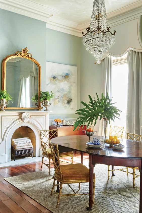 A lovely French chic dining room with light blue walls, a non working fireplace, a stained table and vintage chairs, a large crystal chandelier
