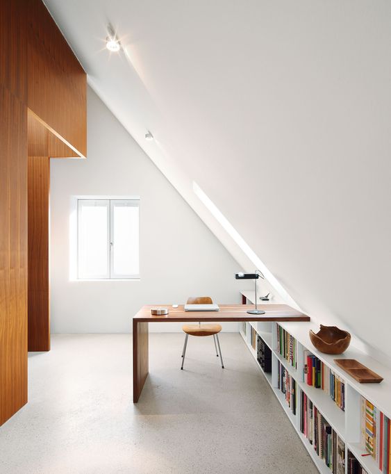 A minimalist attic home office with a long bookshelf and a built in desk, a chic table lamp and a comfy leather chair is filled with light and chic