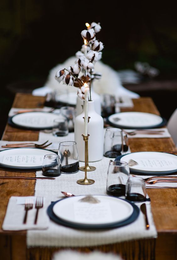a monochromatic winter tablescape with a white runner and placemats, white vases with cotton, black chargers and black bottom glasses plus copper cutlery