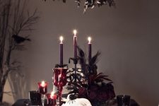 a moody and chic Halloween tablescape with black linens, purple candles and deep red blooms, skulls, crows, spiders and black goblets