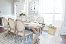 a neutral French country dining room with grey walls, white paneling, a rustic dining table, vintage neutral chairs, a ruffle chair cover on one of them and a crystal chandelier