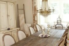 a neutral French dining space with a large storage unit, a stained table, white vintage chairs, a crystal chandelier and some vintage decor