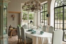 a rustic French dining space with large windows, a stone floor, a vintage buffet, an oval table and neutral chairs
