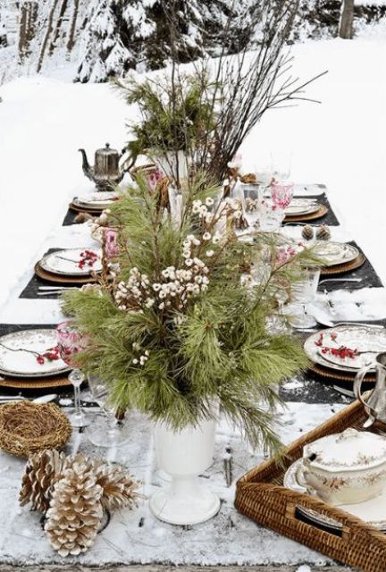 a rustic outdoor winter table with a white runner, an evergreen and berry centerpiece, snowy pinecones, berries for each palce setting