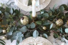 a simple and cute winter table with a greenery and gold ornament runner, white patterned porcelain and greenery and white berries on plates