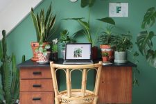 a small and chic attic home office with a green accent wall, a retro desk and a rattan chair, lots of plants and natural light coming through a skylight