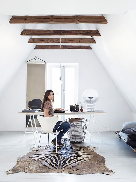 a small attic home office with wooden beams on the ceiling, an airy trestle desk, a white chair, a creative rug, a metal basket and a sofa in the corner