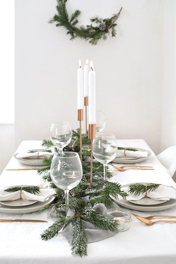 a stylish and fresh winter table with a snowy evergreen and pinecone runner, evergreens, gold cutlery and a matching wreath on the wall