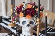 a stylish modern Halloween table setting with bold blooms in a skull, deep toned candles, black and white linens and skeleton hands