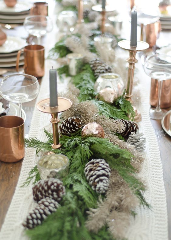 a stylish winter table with a knit runner, evergreen and snowy pinecone runner, grey candles, copper mugs and ornaments