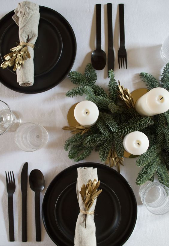 a stylish winter tablescape with evergreens, pillar candles, black plates and chargers and cutlery plus gold branches
