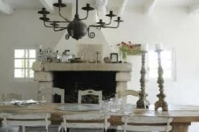 a vintage rustic dining room done in neutrals, with a large hearth, a wooden dining table and chairs, a black metal chandelier and lots of greenery