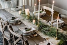 a winter table setting with an evergreen table runner, various candles, bells, neutral porcelain