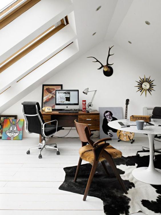 an eclectic attic home office with large skylights, a stained desk and a black leather chair, a round table and a wooden chair, a cool rug and some artworks