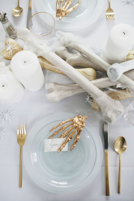 an elegant white and gold Halloween tablescape with bones, white spiders, candles, gold skeleton hands and glass plates