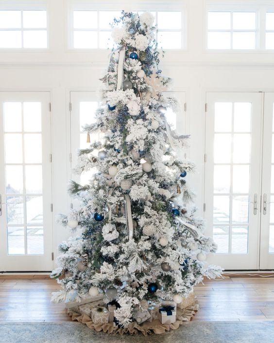 a snowy Christmas tree with white and bold blue ornaments and striped ribbons plus a burlap skirt
