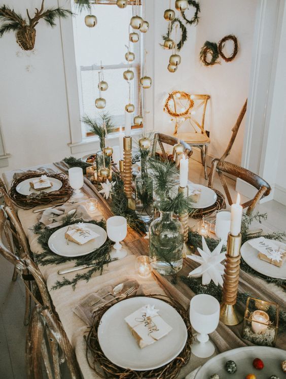 a chic rustic Christmas table with evergreens in bottles and vases, candles, woven placemats and bells hanging over the table