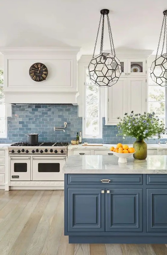 A classic sea inspired kitchen with white shaker cabinets, a blue tile backsplash, a blue kitchen island with white stone countertops and beautiful faceted lamps