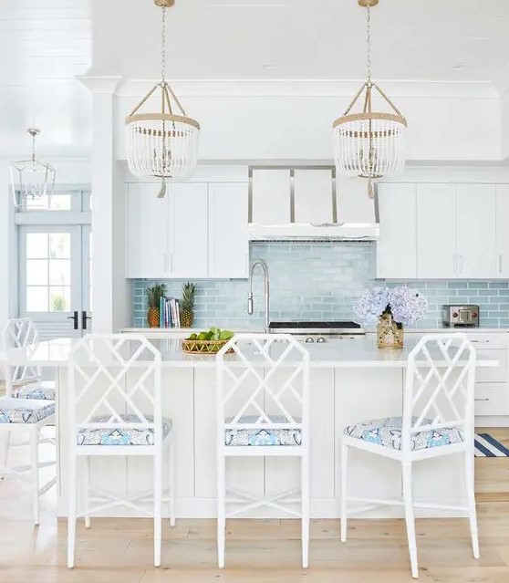 a coastal kitchen with white cabients, a large kitchen island, chic beaded chandeliers, white chairs with printed upholstery