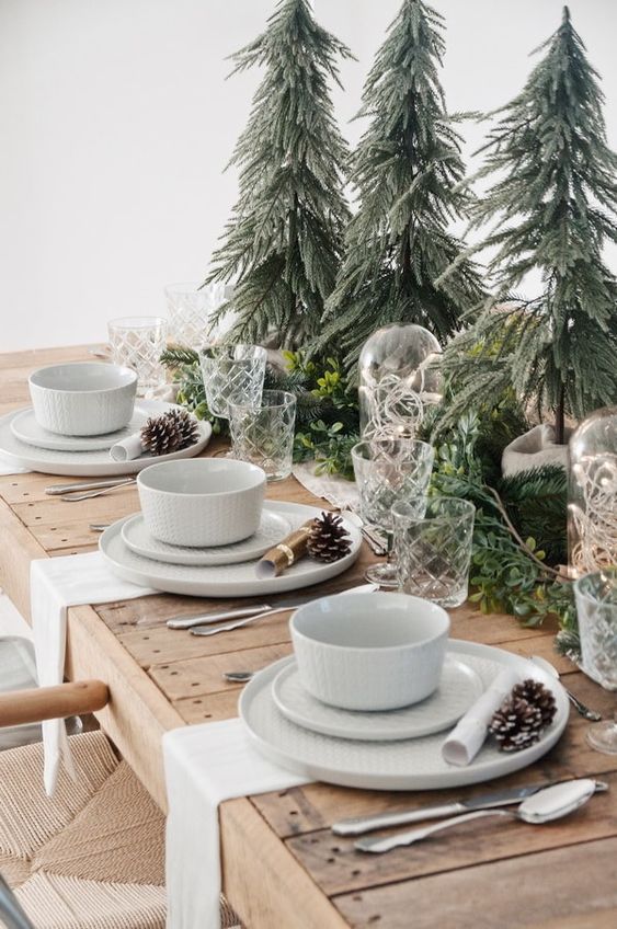 a cozy Christmas tablescape with mini trees, evergreens and greenery, white porcelain and pinecones plus white napkins