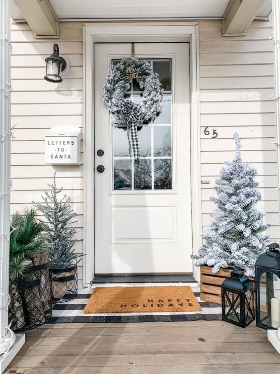 a farmhouse Christmas porch with snowy Christmas trees, candle lanterns, a snowy Christmas wreath is a veyr cozy space