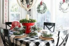 a farmhouse Christmas table with a plaid tablecloth, woven placemats, evergreens, a red stand with greenery and a pillar candle