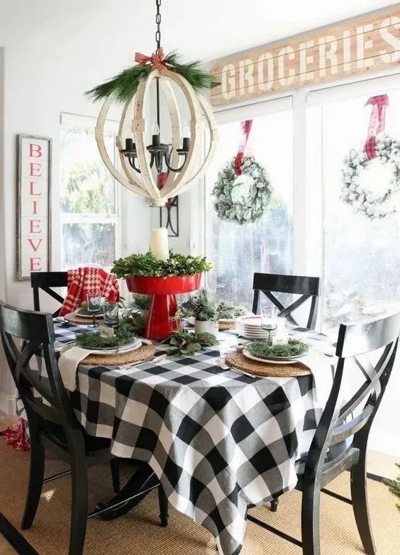 a farmhouse Christmas table with a plaid tablecloth, woven placemats, evergreens, a red stand with greenery and a pillar candle