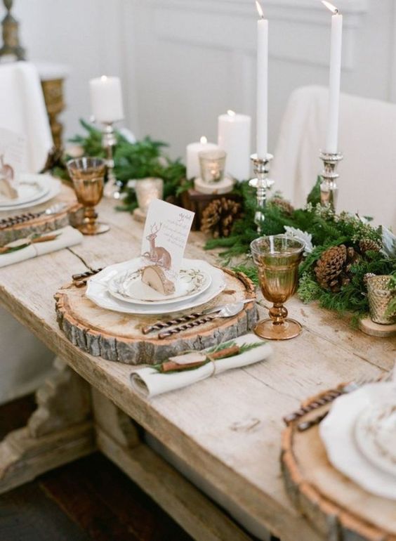 a gorgeous rustic Christmas table with an evergreen and pinecone runner, amber glasses, wood slice placemats, tall candles and deer cards
