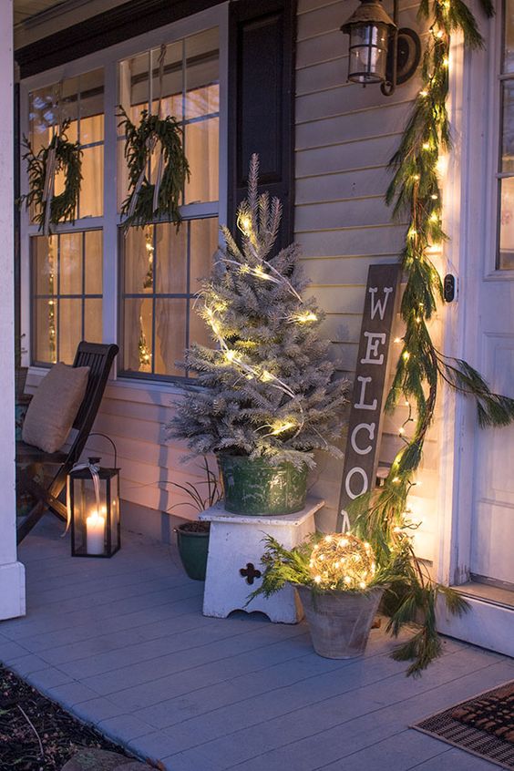 a neutral farmhouse porch with evergreen garlands, wreaths, lights, a snowy Christmas tree and candle lanterns is cozy and inviting