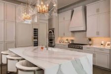 a refined neutral-colored kitchen with a jaw-dropping kitchen island with a waterfall countertop and stunning gold and sheer glass chandeliers