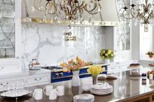 a refined vintage-inspired kitchen in white, with a bold blue cooker, a white marble backsplash, crystal chandeliers and a vintage desk as a kitchen island