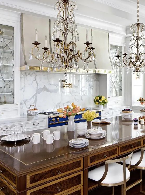 A refined vintage inspired kitchen in white, with a bold blue cooker, a white marble backsplash, crystal chandeliers and a vintage desk as a kitchen island