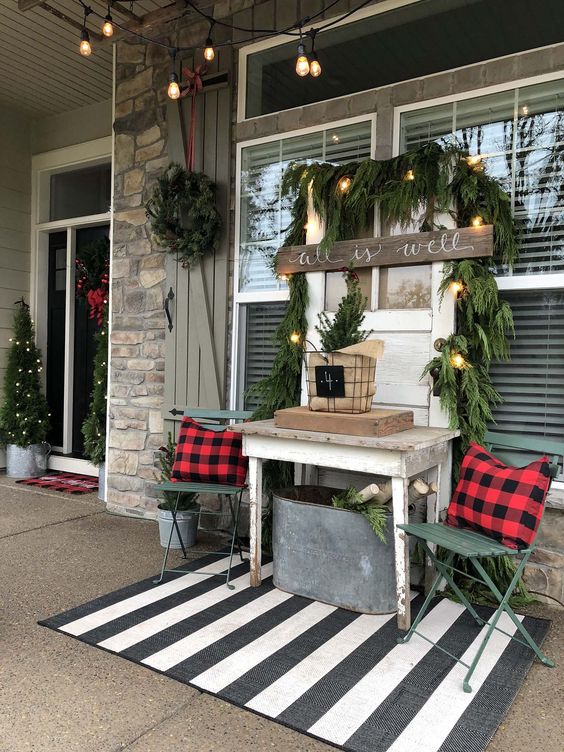 a rustic Christmas porch with evergreens, plaid pillows, a bucket with firewood, lights, a sign and trees framing the doorway