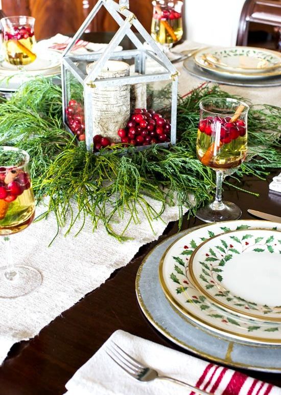 a rustic Christmas table setting with a fabric runner, evergreens, a lantern with berries and a stump candleholder plus printed porcelain