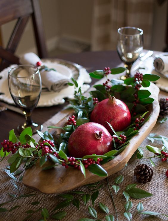 a rustic Christmas table with a burlap runner, greenery, berries, pinecones and a dough bowl with pomegranates and berries