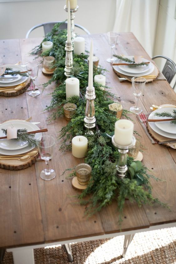 a stylish rustic Christmas table with an evergreen runner, candles, wooden placemats, cinnamon sticks and gold cutlery