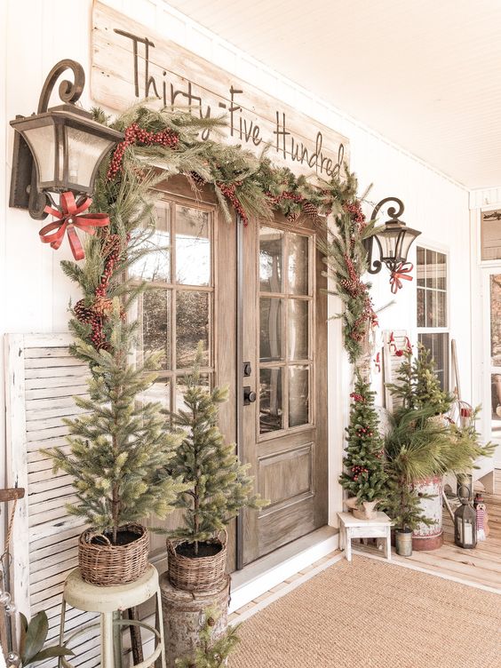 a vintage rustic Christmas porch with an evergreen garland with berries and pinecones, mini trees that aren’t decorated and lanterns