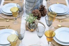 a whimsy rustic Christmas table with wooden placemats, amber and grey glasses, pinecones, antlers, potted greenery and metal candle holders