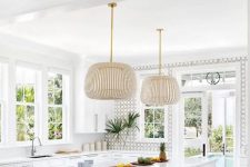 an airy and vivacious neutral beach kitchen with white shaker cabinets, an accent wall, a kitchen island with a dining space and catchy pendant lamps