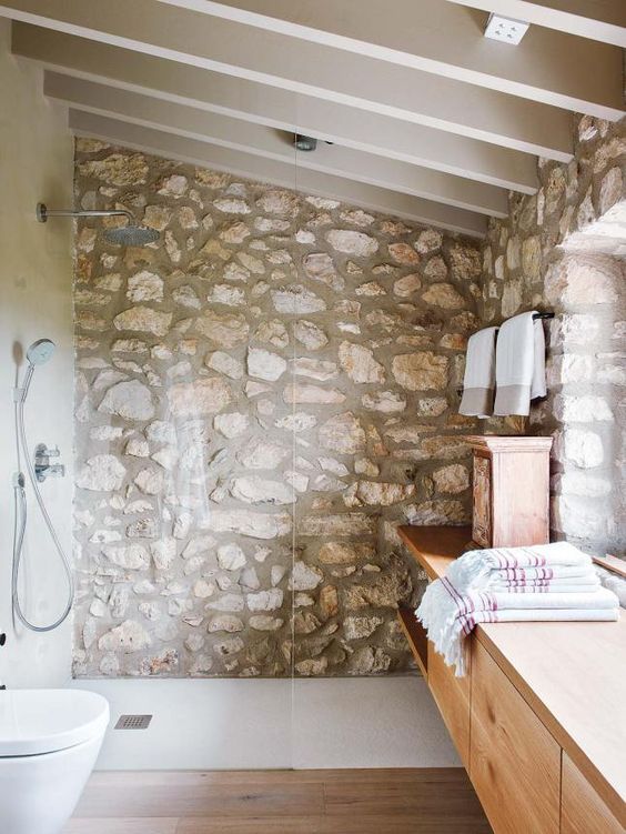a chic attic bathroom with a rough stone wall, a sleke wooden vanity and enough natural light incoming