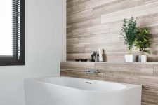 a clean contemporary bathroom done with laminate, with a chic bathtub and a raised shelf over it, with a window and a printed rug