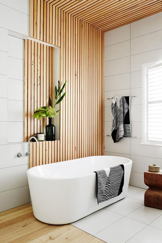a contemporary bathroom clad with large scale white tiles and wooden slabs, with a wooden floor, a chic tub and some lovely decor