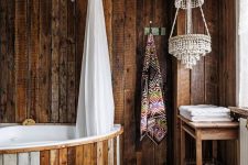 a creative bathroom clad with dark stained rough wood, with a bathtub clad with wood, beaded chandeliers and printed textiles